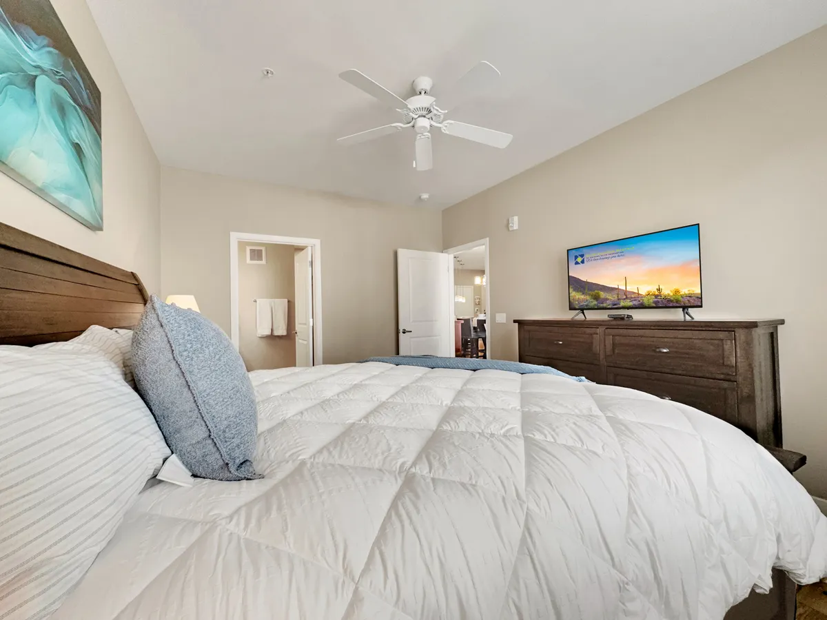 A serene bedroom featuring a bed with a white comforter, a wooden dresser, a wall-mounted TV, and tasteful artwork on the wall. Soft natural light brightens the room, and the overall decor feels inviting and restful.
