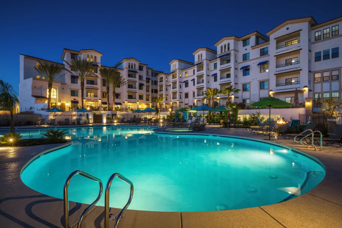 A sparkling illuminated swimming pool framed by swaying palm trees, with balconies and lounge chairs visible. The evening setting invites relaxation and socializing by the water in a resort-like atmosphere.