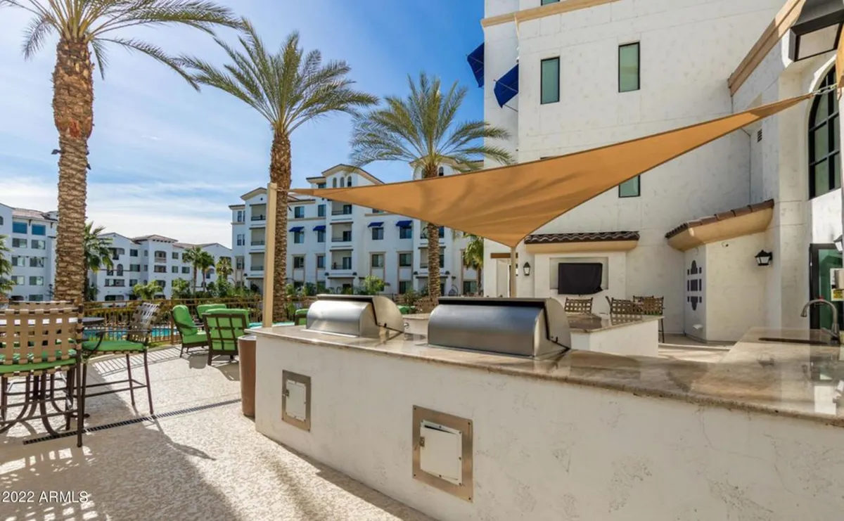 A communal outdoor grill area with comfortable seating, palm trees, and residential buildings in the background. It captures a sense of community, leisure, and the potential for evening gatherings or celebrations by the pool.
