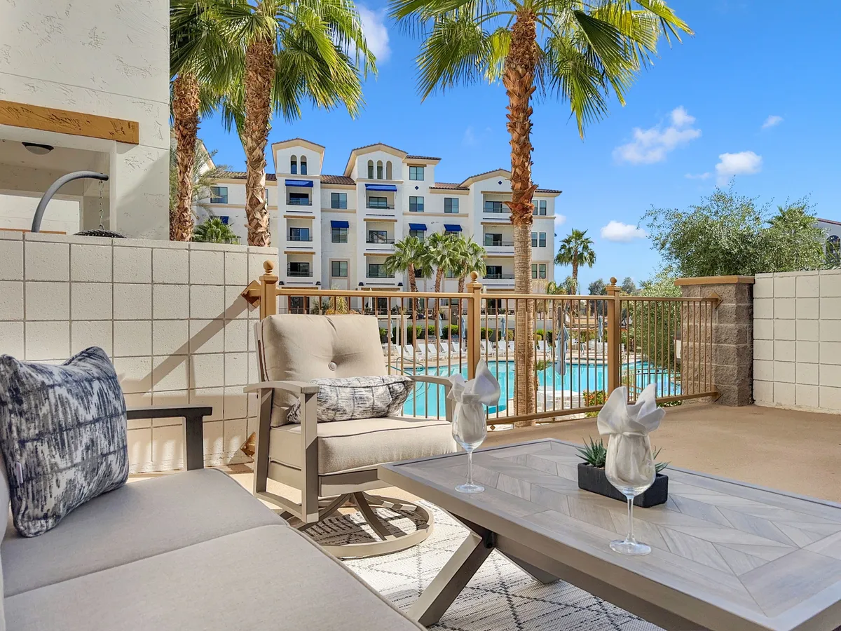 A sunlit outdoor seating area surrounds a sparkling swimming pool, with lush palm trees and the contemporary building facade in the background. Comfortable lounge chairs and shaded tables suggest spaces for relaxation, morning coffee, or casual socializing.