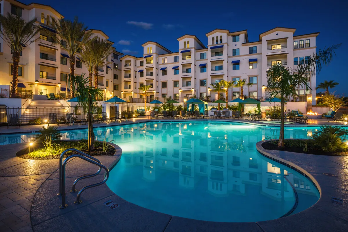 A stunning exterior shot of the resort-style swimming pool surrounded by palm trees, illuminated building facade, lounge chairs, and canopy-covered seating areas. The pool area exudes a calm, luxurious ambiance ideal for relaxation and social gatherings.