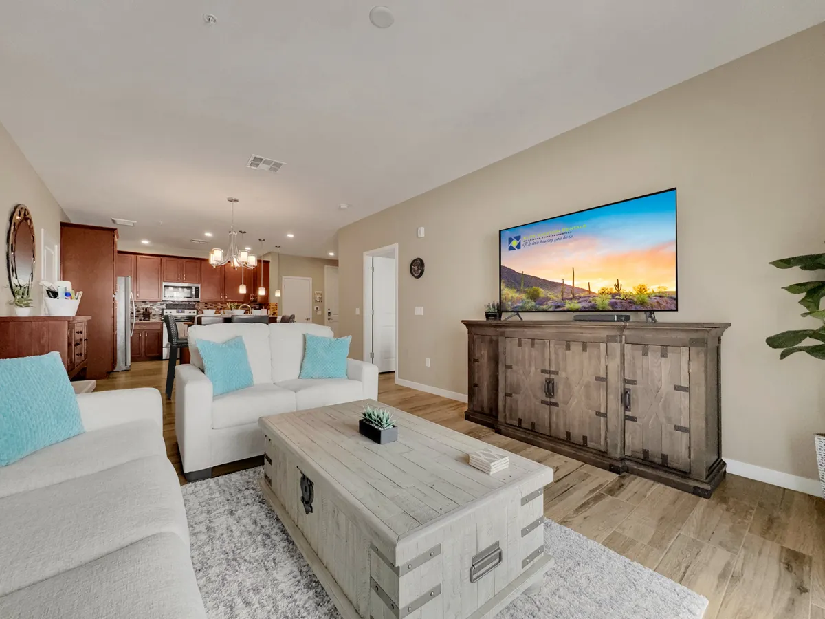 This image captures a cozy, open-concept living area featuring a plush sofa with turquoise cushions, a wooden coffee table, and a television displaying a scenic image. The kitchen area is visible in the background, reinforcing the flow between living and dining spaces.